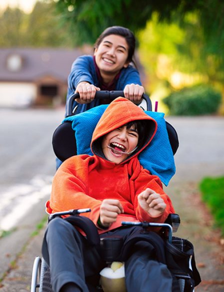 Big sister pushing disabled little brother in wheelchair around neighborhood, laughing and smiling